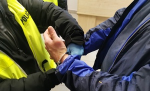 Policjant w ubraniu cywilnym w kamizelce z napisem policja zakłada zatrzymanemu kajdanki