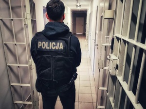 Policjant w ubraniu cywilnym w kamizelce zx napisem Policja stoi na korytarzu pomieszczenia dla osób zatrzymanych.