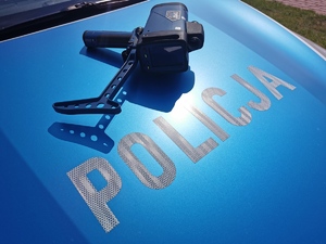 Ręczny miernik prędkości leżący na pokrywie silnika radiowozu, nad napisem policja.