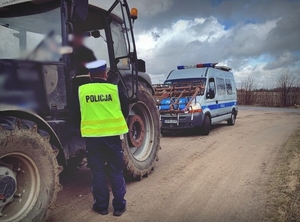 Policjant ruchu drogowego kontroluje ciągnik rolniczy. Za maszyną widać stojący na poboczu radiowóz policyjny