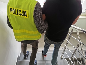 Policjant w ubraniu cywilnym, w kamizelce z napisem policja prowadzi zatrzymanego mężczyznę po schodach komendy.