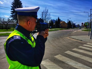 Umundurowany policjant stoi przy jezdni i mierzy prędkość pojazdom.