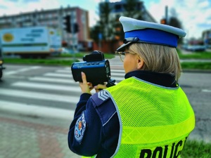 Umundurowana policjantka stoi przy jezdni i mierzy prędkość pojazdom.