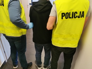 Dwóch policjantów w ubraniach cywilnych w kamizelkach z napisem policja trzymają zatrzymanego mężczyznę, który ma założone kajdanki na ręce trzymane z przodu.