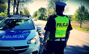 Policjant ruchu drogowego (w czapce z białym otokiem i kamizelce z napisem POLICJA) przy radiowozie patrzy się w stronę drogi po której jedzie samochód osobowy