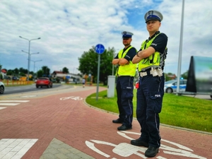 Dwóch umundurowanych policjantów stojących przy przejściu dla pieszych i ścieżce dla rowerów.