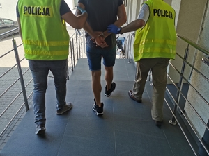 Dwóch policjantów w ubraniach cywilnym z kamizelkami z napisem policja trzymają zatrzymanego, który ma założone kajdanki na ręce trzymane z tyłu, prowadzą go schodami zewnętrznymi komendy