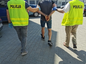 Dwóch policjantów w ubraniach cywilnym z kamizelkami z napisem policja trzymają zatrzymanego, który ma założone kajdanki na ręce trzymane z tyłu. Policjanci prowadzą go dziedzińcem komendy.