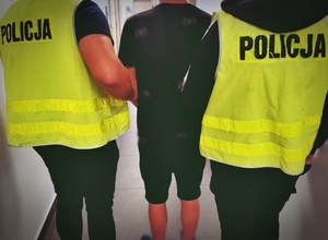 Mężczyzna z rękoma trzymanymi z przodu zakutymi w kajdanki, Mężczyzna trzymany jest przez dwóch funkcjonariuszy w cywilnym ubraniu w kamizelkach odblaskowych z napisem POLICJA