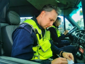 Policjanci siedzą w radiowozie i piszą w notatnikach służbowych