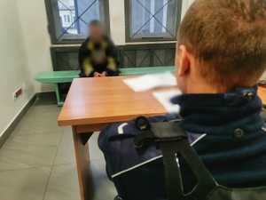 Zatrzymany poszukiwany mężczyzna siedzi na ławce w pomieszczeniu dla osób zatrzymanych, ma założone kajdanki na ręce trzymane z przodu. Przed nim, przy biurku siedzi policjant.