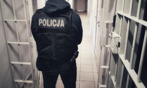 Policjant w ubraniu cywilnym, w kamizelce z napisem policja , stoi w pomieszczeniu dla osób zatrzymanych.
