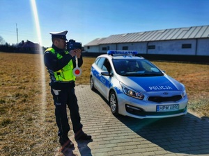 Policjant stoi przy radiowozie i mierzy prędkość pojazdom