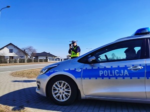 Policjant stoi przy radiowozie i mierzy prędkość pojazdom