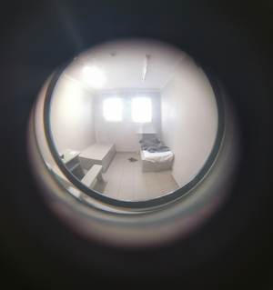 Obraz z judasza w drzwiach celi. Po lewej stół i ławka po prawej na łóżku leży mężczyzna przykryty kocem