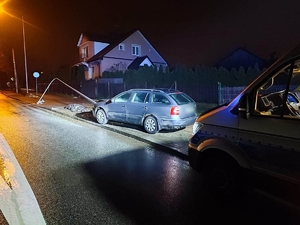 Radiowóz oznakowany z włączonymi światłami błyskowymi stoi na chodniku, a przed nim samochód osobowy, który uderzył w latarnię.