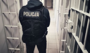 Policjant w ubraniu cywilnym, w kamizelce z napisem policja stoi na korytarzu policyjnego aresztu.