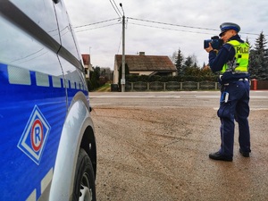 Policjant stoi przy jezdni i mierzy prędkość jadącym pojazdom.