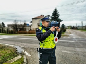Policjant stoi przy jezdni i mierzy prędkość jadącym pojazdom.