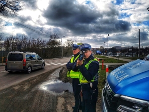 Dwóch policjantów stoi przy przejściu dla pieszych. Jeden z nich mierzy prędkość jadącym pojazdom.
