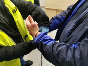 Policjant w ubraniu cywilnym, w kamizelce z napisem policja zakłada kajdanki zatrzymanemu mężczyźnie