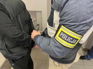 Policjant w ubraniu cywilnym z opaską na ręku z napisem policja zakłada kajdanki zatrzymanemu mężczyźnie