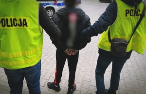 Dwóch policjantów w ubraniach cywilnych, w kamizelkach z napisem policja trzymają podejrzanego mężczyznę, który ma założone kajdanki na ręce trzymane z tyłu.