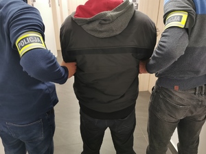 Dwóch policjantów w ubraniach cywilnych, z opaskami na ręku z napisem policja trzymają zatrzymanego mężczyznę.