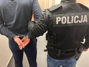 Policjant w kamizelce z napisem policja trzyma zatrzymanego mężczyznę, który ma założone kajdanki na ręce trzymane z tyłu