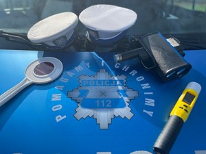 Dwie czapki policyjne na pokrywie silnika radiowozu, obok ręczny miernik prędkości, alcoblow oraz tarcza do zatrzymywania pojazdów.
