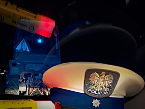 czapka policyjna i urządzenie do pomiaru alkoholu. W oknie radiowozu odbija się ciągnik rolniczy i czerwona lampka urządzenia do pomiaru alkoholu